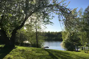 Ruhiger See im Campingbereich, umgeben von Bäumen und grünem Gras, mit Booten am Ufer.
