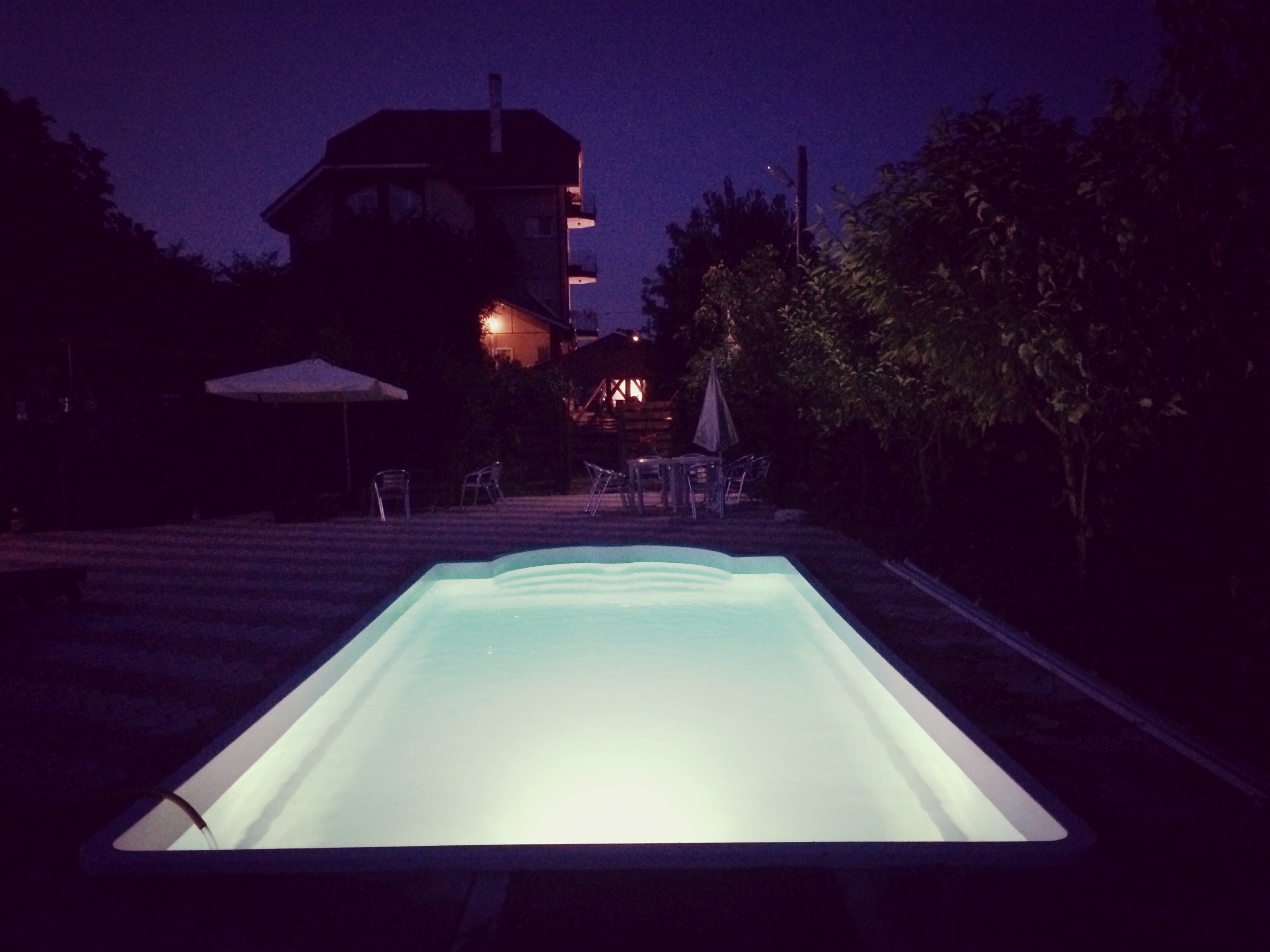 Beleuchteter Camping-Pool am Abend mit Liegen, Sonnenschirm und Wegen neben Bäumen und einem Gebäude.