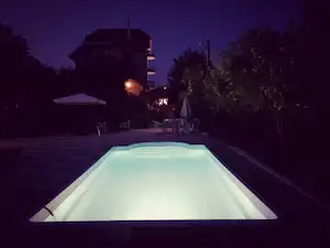 Beleuchteter Camping-Pool am Abend mit Liegen, Sonnenschirm und Wegen neben Bäumen und einem Gebäude.