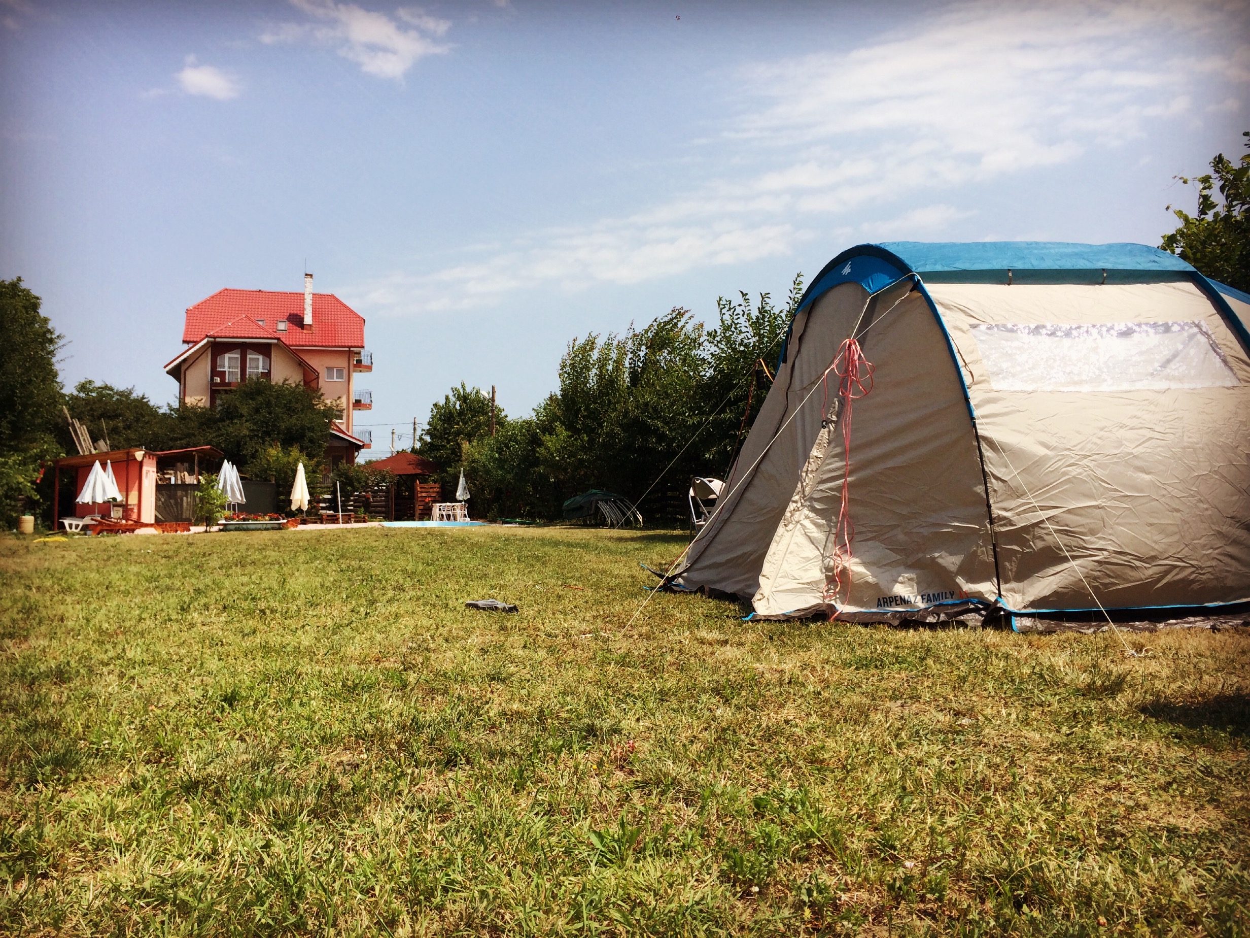 Campingzelt auf grünem Rasen, im Hintergrund ein Gebäude mit rotem Dach und Bäumen am Campingplatz.
