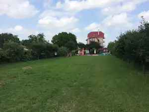 Grüner Campingrasen mit Haus und Terrasse im Hintergrund, umgeben von Bäumen und Hecken.