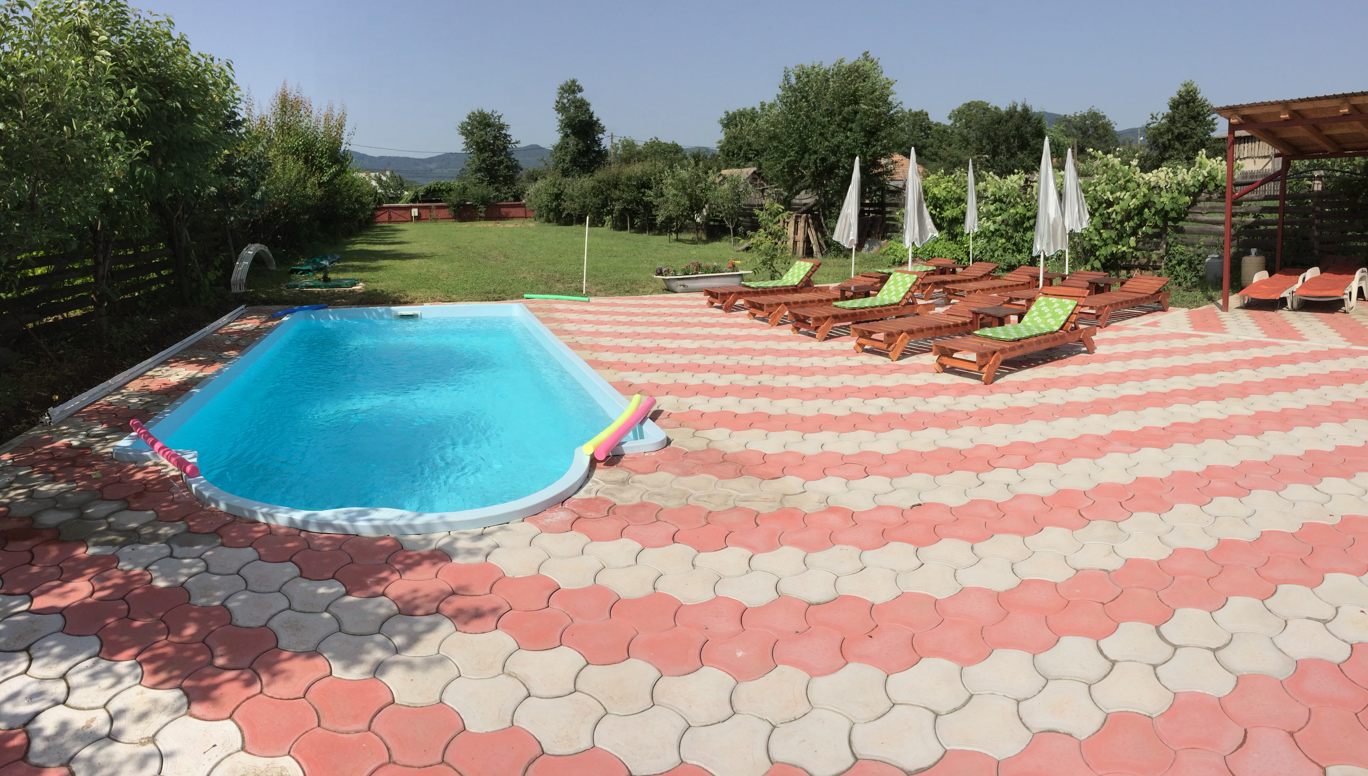Blauer Pool mit gepflasterter Fläche und Sonnenliegen im Garten, umgeben von Bäumen und Hecken.