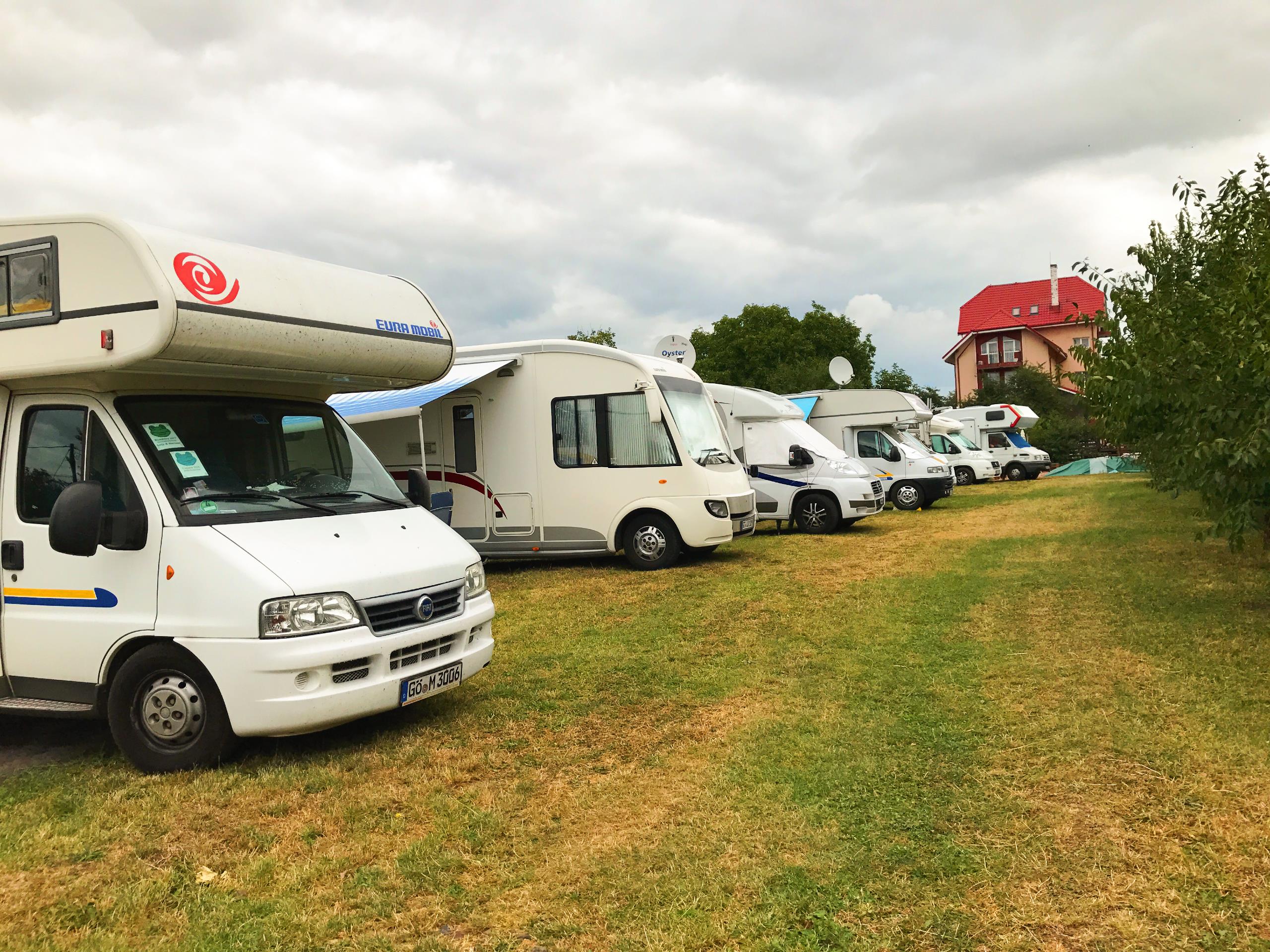 Mehrere Wohnmobile stehen auf einer Campingwiese, im Hintergrund ein rotes Haus und weitere Fahrzeuge.