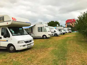Mehrere Wohnmobile stehen auf einer Campingwiese, im Hintergrund ein rotes Haus und weitere Fahrzeuge.