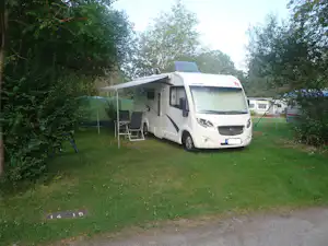 Wohnmobil auf einem Campingplatz mit Vorzelt, zwei Stühlen und Tisch auf dem Gras, umgeben von Bäumen.