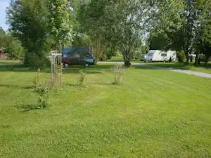 Campingwiese mit Wohnwagen und Auto am Stellplatz, umgeben von Bäumen und einem gepflegten Weg.