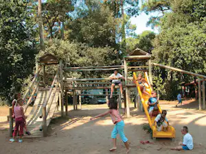 Spielplatz auf dem Campingplatz mit Klettergerüst, Rutsche und Kindern im Grünen zwischen Bäumen.