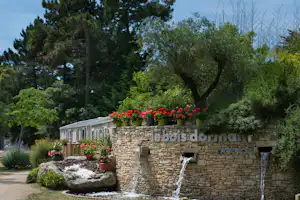 Steinmauer mit Springbrunnen und Blumenbeeten, davor ein Weg und Bäume im Hintergrund.