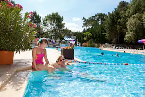 Familie im Camping-Schwimmbad: Kinder und Erwachsene im Wasser, Sonnenschirme, Liegen und Bäume im Hintergrund.