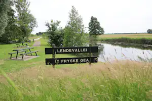 Holzbänke und ein Schild am Ufer eines ruhigen Sees, umgeben von Gras und Bäumen in einer Campinganlage.