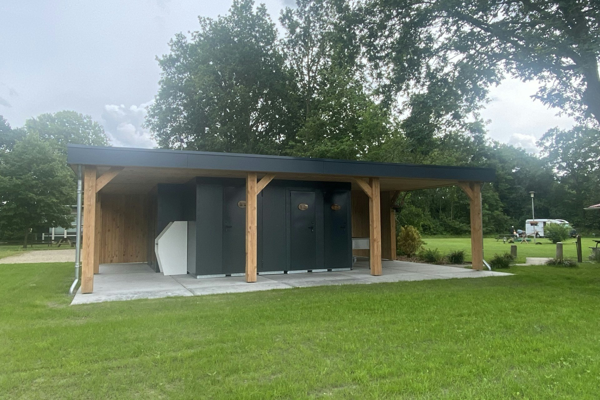 Moderner Stellplatz mit überdachtem Holzcarport und Vorzelt, daneben Rasen und Bäume im Campinggelände.