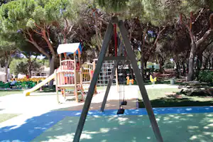 Spielplatz auf dem Campingplatz mit Rutsche, Klettergerüst und Schaukeln auf gepflastertem Boden im Schatten der Bäume.