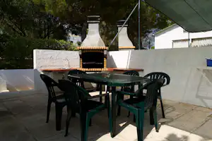 Grillplatz mit Tisch und Stühlen auf gepflasterter Terrasse, gemauerter Grill im Hintergrund und Zeltplane am Rand.