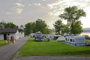 Campingplatz mit Wohnwagen und Zelten auf dem Stellplatz, Rasenfläche, Bäume und See im Hintergrund.