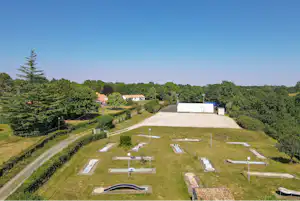 Campingplatz mit Stellplätzen auf einer grünen Wiese, weißen Markierungen und Wegen, im Hintergrund Bäume und Gebäude.