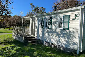 Weiße Wohnwagen-Unterkunft mit Veranda und Stufen, umgeben von Rasen und Bäumen im Campingbereich.