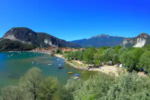 Blick auf den See mit Strand, Booten und Liegewiese am Campingplatz, umgeben von Bäumen und Bergen.