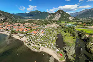 Campingplatz am Fluss mit Stellplätzen, Wohnwagen und Grünflächen, umgeben von Bergen und einem Tal unter blauem Himmel.