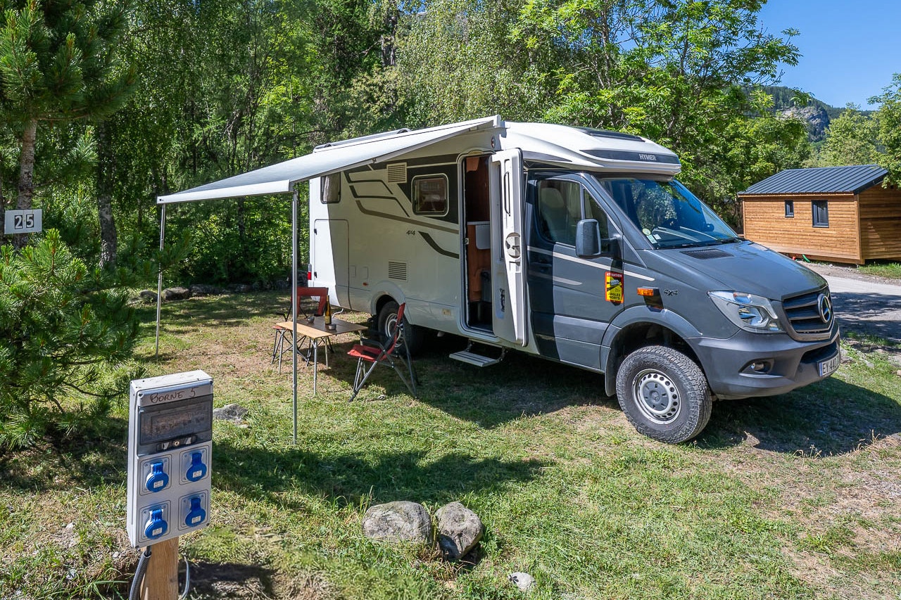 Wohnmobil mit geöffnetem Seitenfenster auf einem Stellplatz im Grünen, neben Sitzbank und Bäumen vor einem Holzhaus.