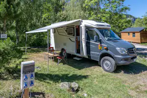 Wohnmobil mit geöffnetem Seitenfenster auf einem Stellplatz im Grünen, neben Sitzbank und Bäumen vor einem Holzhaus.