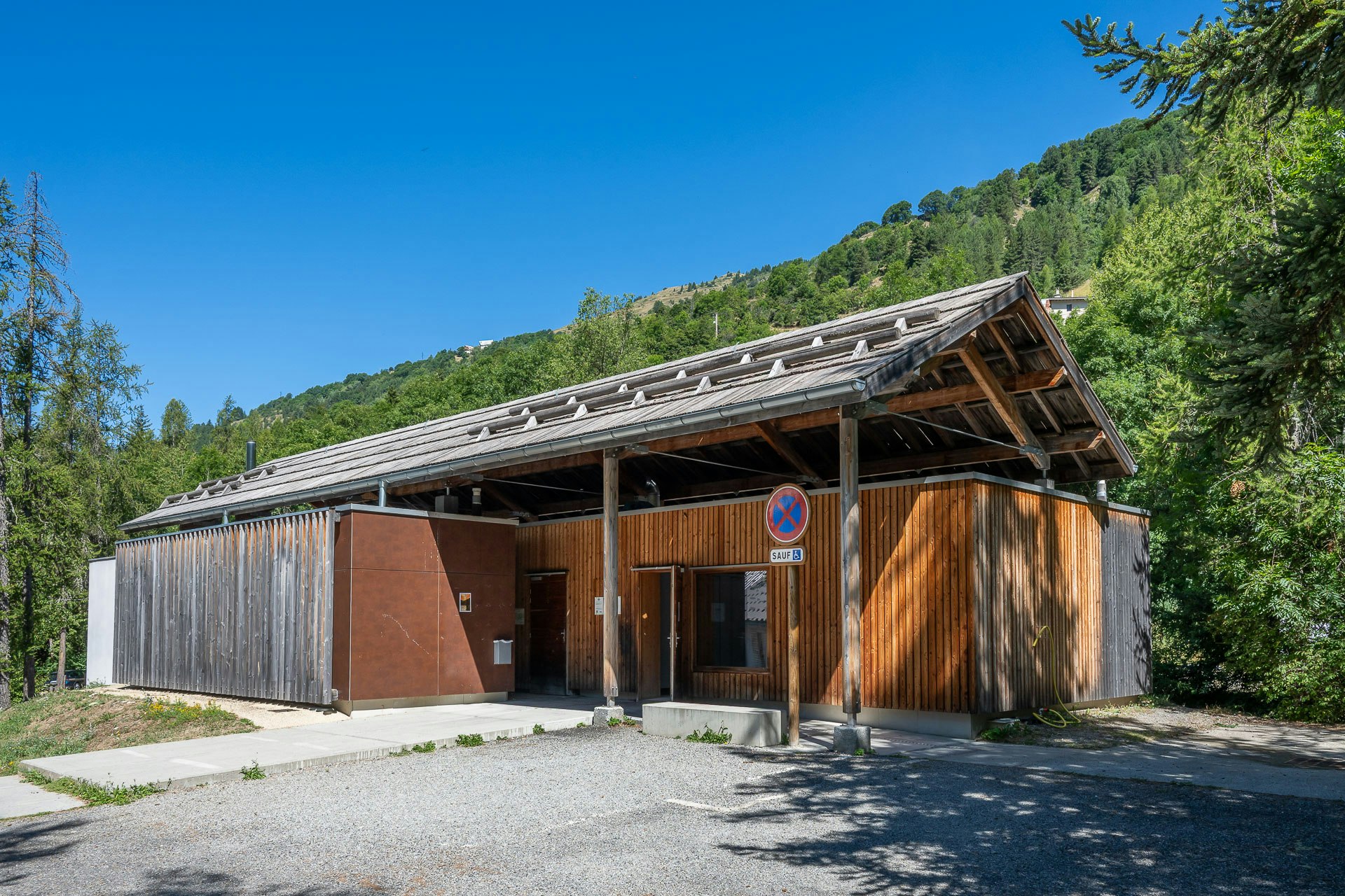 Holzhütte mit überdachtem Eingang und Holzverkleidung, vor einem Kiesweg, umgeben von Bäumen und grünen Hügeln.