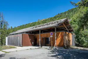 Holzhütte mit überdachtem Eingang und Holzverkleidung, vor einem Kiesweg, umgeben von Bäumen und grünen Hügeln.