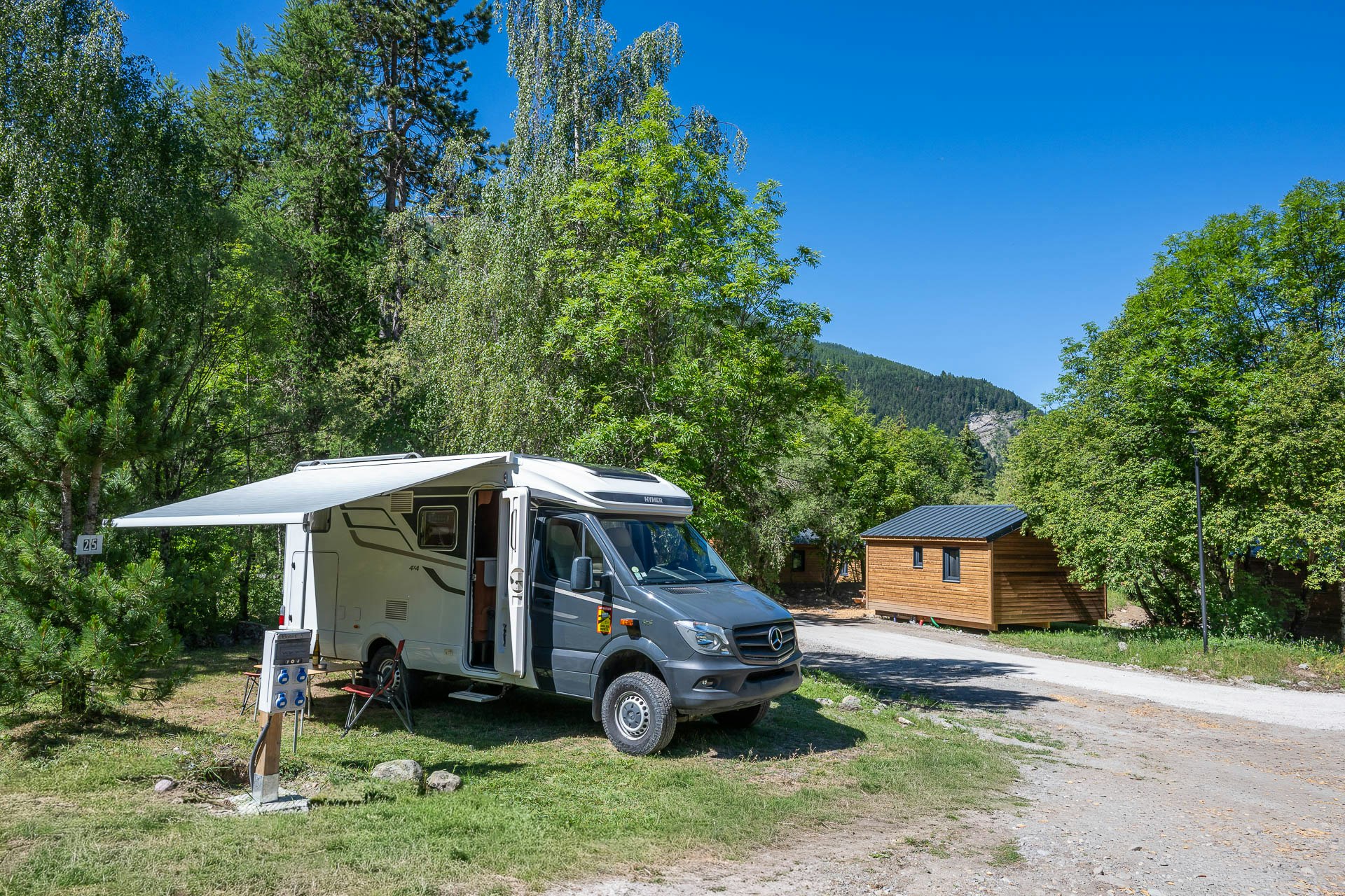 Wohnmobil mit Vorzelt am Stellplatz, neben einem Holzhaus und einem Weg im Grünen, umgeben von Bäumen und Bergen.