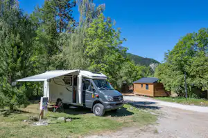 Wohnmobil mit Vorzelt am Stellplatz, neben einem Holzhaus und einem Weg im Grünen, umgeben von Bäumen und Bergen.