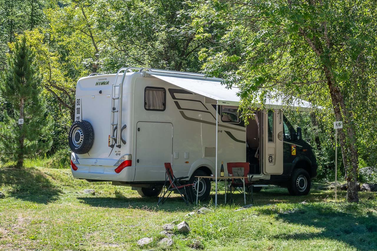 Wohnmobil auf einem Campingplatz am Hang, neben Bäumen und Wiese, im Hintergrund hohe Berge mit schneebedeckten Gipfeln.