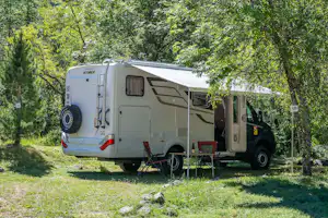 Wohnmobil auf einem Campingplatz am Hang, neben Bäumen und Wiese, im Hintergrund hohe Berge mit schneebedeckten Gipfeln.