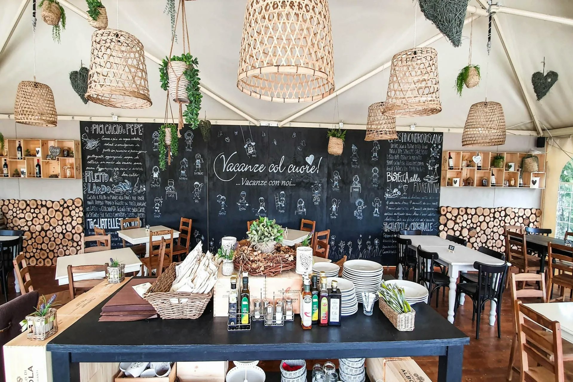 Innenansicht eines Campingzelts mit Bar-Tisch, Holzstühlen, Regalen mit Flaschen und einer Tafel mit handschriftlichen Texten.