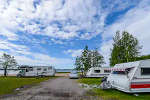 Wohnmobile und Auto auf einem Campingplatz am See, Bäume am Ufer und großer Himmel mit Wolken.