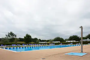 Blaues Schwimmbecken mit Liegen am Campingplatz, im Hintergrund Wohnwagen und Bäume unter bewölktem Himmel.