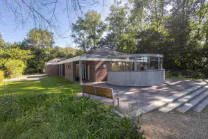 Campinggebäude mit Terrasse, Ziegelwand und Glasfront, gepflasterter Weg und Bank im grünen Garten mit Bäumen.