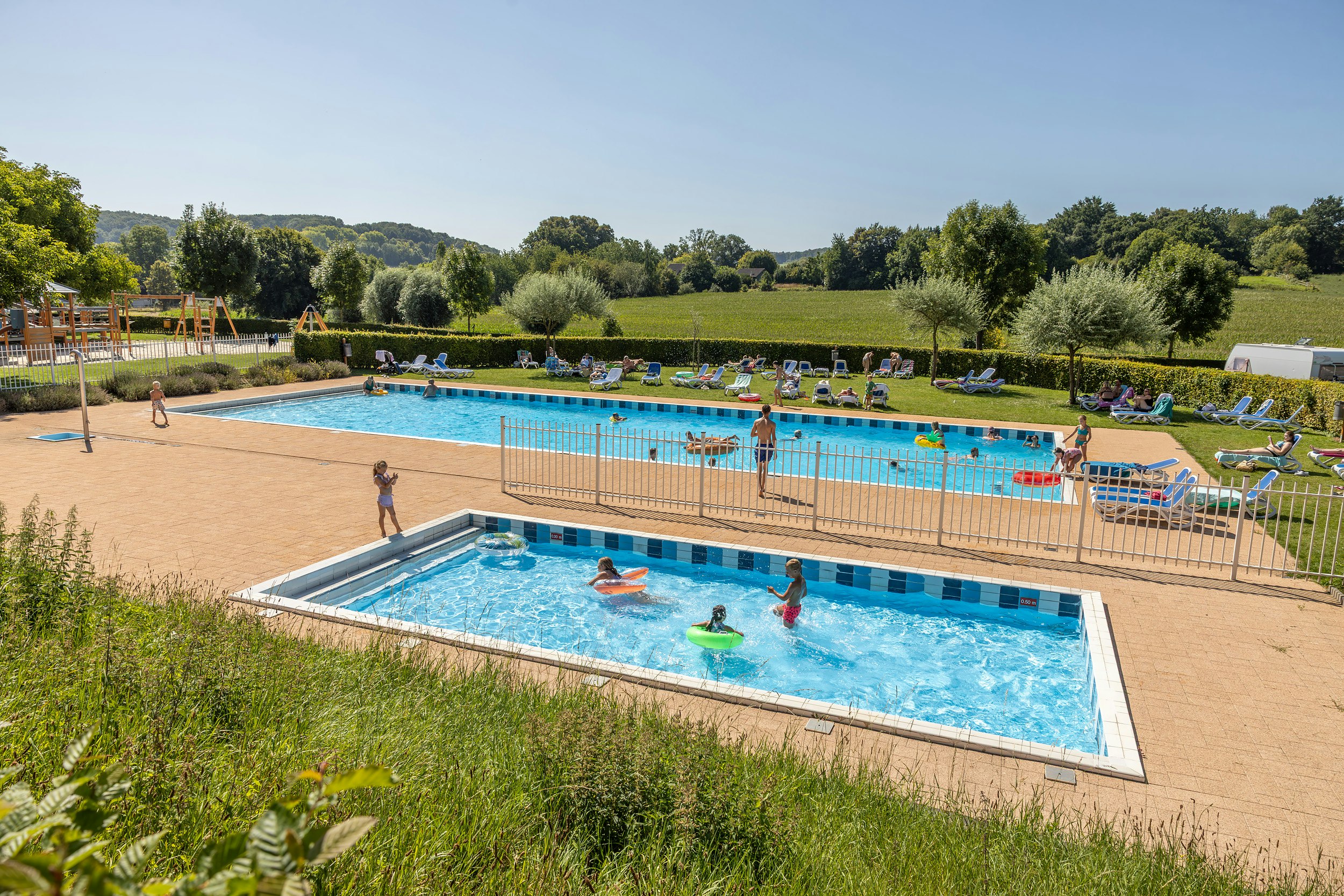 Schwimmbecken auf dem Campingplatz mit Liegestühlen, Sonnenschirmen und gepflegtem Garten, im Hintergrund Bäume und Wiesen.