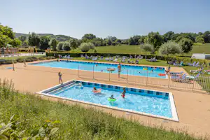 Schwimmbecken auf dem Campingplatz mit Liegestühlen, Sonnenschirmen und gepflegtem Garten, im Hintergrund Bäume und Wiesen.