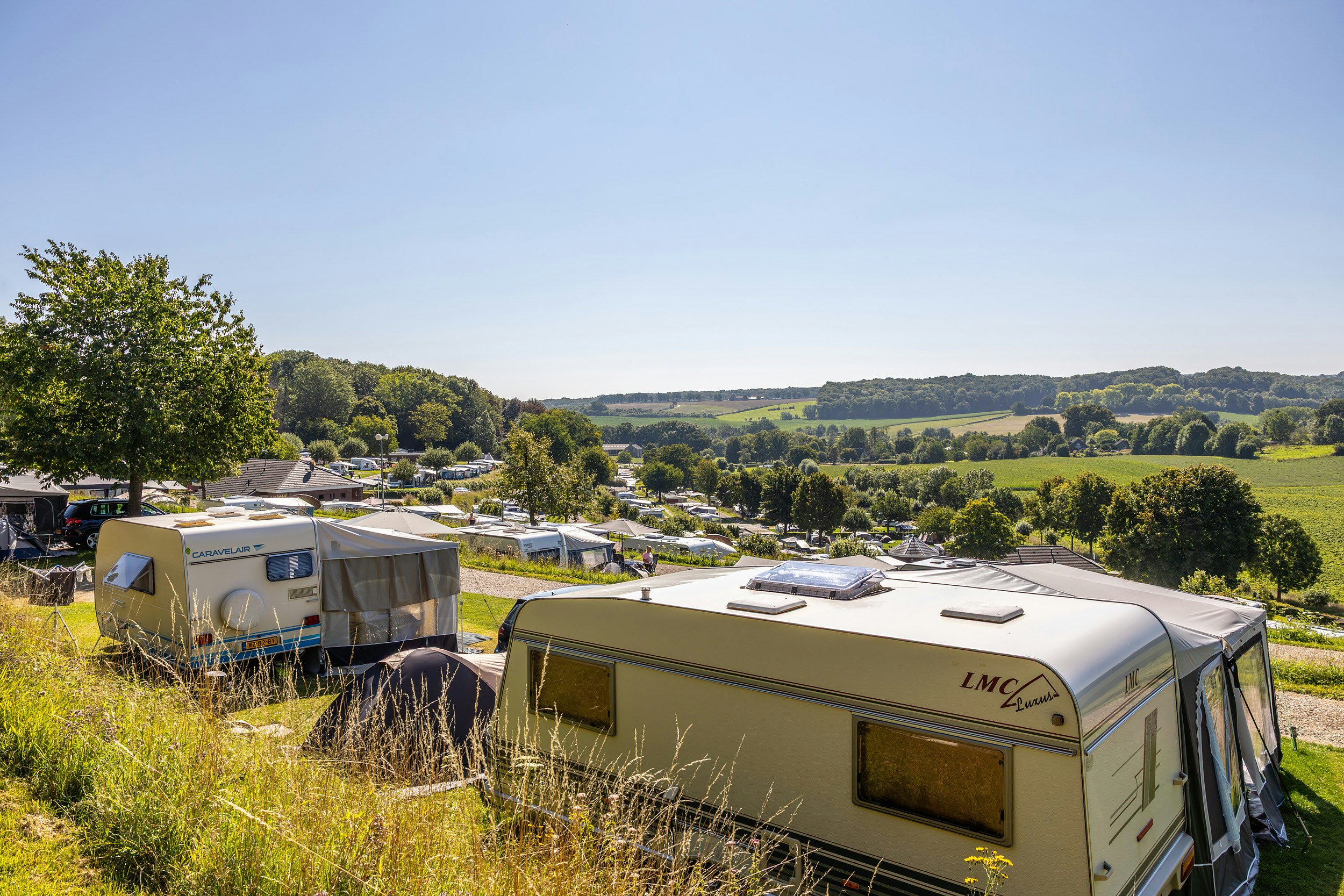 Wohnwagen auf dem Campingplatz mit weiteren Caravans, Bäumen und Blick über grüne Hügel in der Ferne.