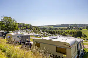 Wohnwagen auf dem Campingplatz mit weiteren Caravans, Bäumen und Blick über grüne Hügel in der Ferne.
