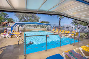 Beheizter Camping-Pool unter einem überdachten Bereich mit Liegen, Sonnenschirmen und Bäumen im Hintergrund.