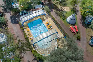 Luftaufnahme: beheizter Camping-Pool mit Liegestühlen, daneben Wohnwagen und Zelte, umgeben von Bäumen und Wegen.