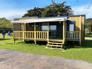 Modernes Campinghaus mit Holzterrasse, großen Fenstern und Markise auf einem gepflegten Rasen, im Hintergrund Bäume und Zaun.