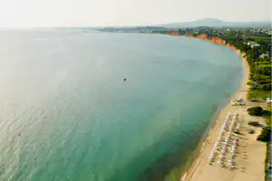 Luftaufnahme eines langen Strands am Meer mit Liegen, Sonnenschirmen und Spazierweg entlang der Küste.