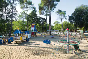 Spielplatz am Campingplatz mit Sandboden, Klettergerüst, Rutsche, Wippe und Trampolin vor hohen Bäumen.