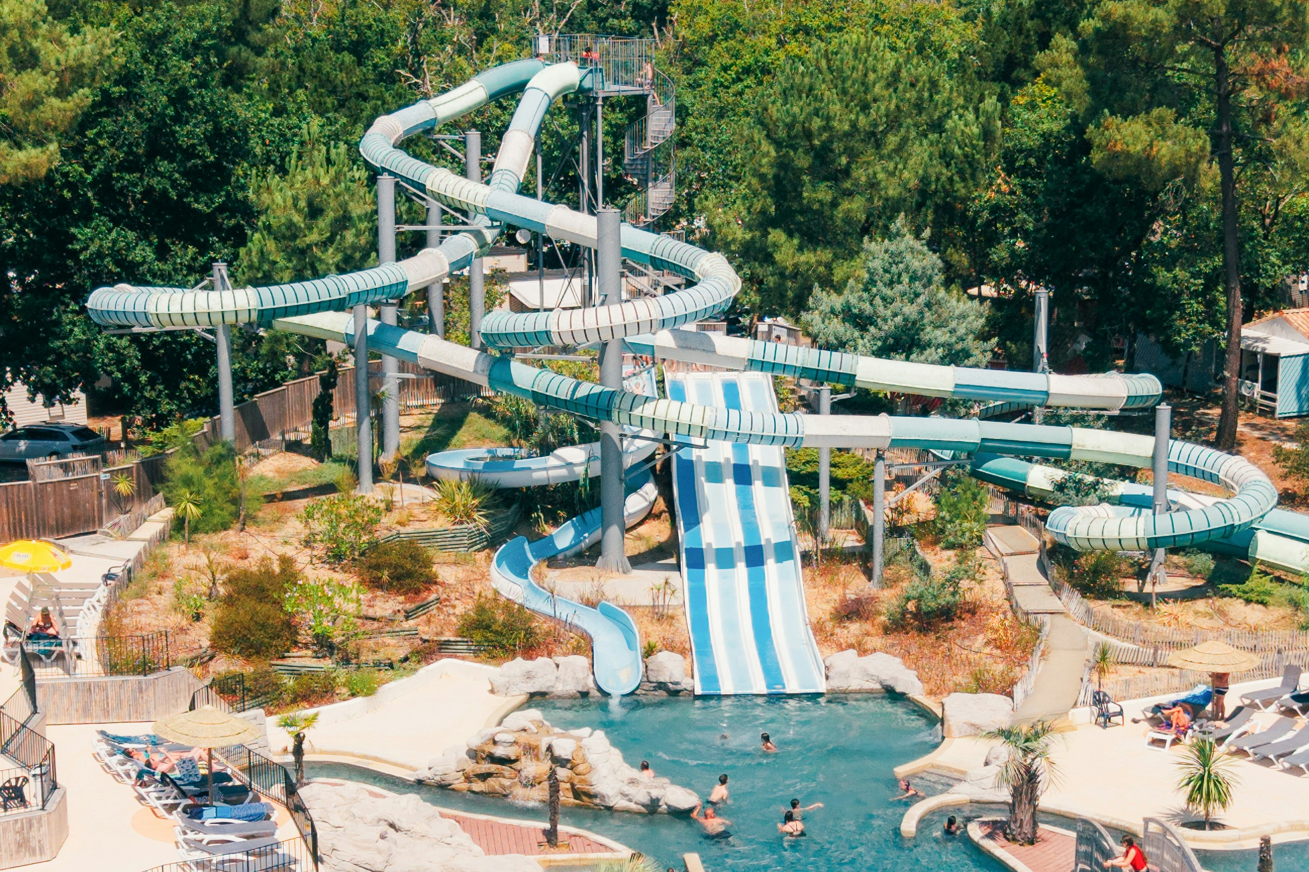 Wasserrutschen und ein türkisfarbenes Schwimmbecken in einem Campingpark, umgeben von Bäumen und Liegen.