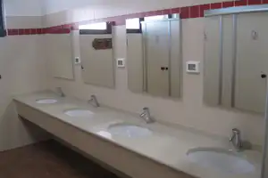 Waschraum mit mehreren Waschbecken, Spiegelfächern und Armaturen, daneben Kabinentüren und Fliesenwände.