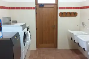 Waschraum mit Holztür, Waschmaschine, Waschbecken und Ablage, weiße Fliesenwand mit roten Streifen.