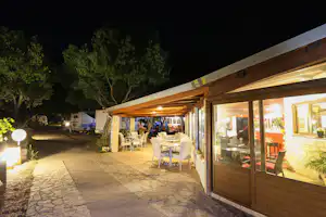 Beleuchtete Camping-Terrasse in der Nacht mit Tischen und Stühlen vor einem Gebäude, im Hintergrund Zelte und Bäume.