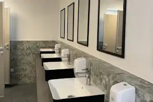 Modernes Badezimmer mit mehreren Waschbecken, Spiegeln und grauen Fliesen an der Wand, daneben eine Toilette.