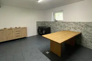Innenraum mit Holztisch, grauem Boden, Kamin und Fenster im Hintergrund, dazu eine Holzkommode an der Wand.