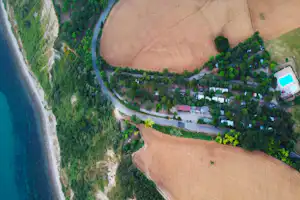 Luftaufnahme eines Campingplatzes am Fluss mit Wegen, Wohnwagen und Zelten im Wald, nahe einem großen Feld.